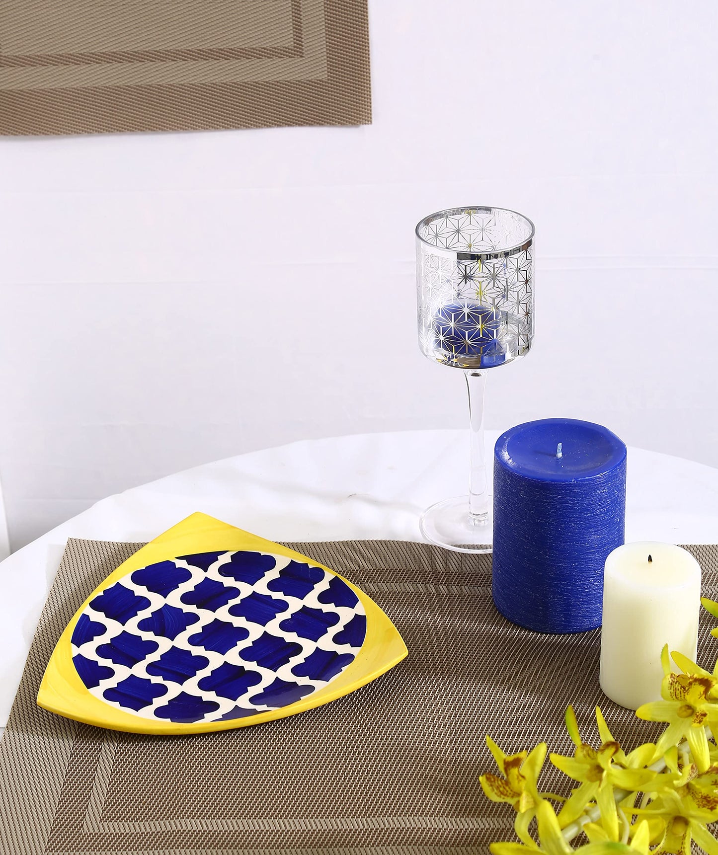 A triangle-shaped, blue and yellow patterned ceramic plate placed on a table with a glass vase and a blue candle holder in the background.