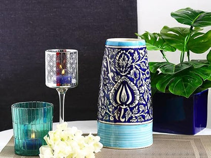 A handmade blue ceramic vase with a floral Mughal pattern, a candle, and a green plant beside it on a table.