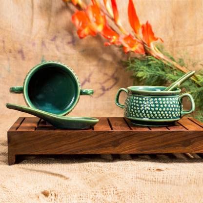 A set of two green ceramic handmade double handled soup bowls with a bubble design, accompanied by a matching green spoon, displayed on a wooden tray against a natural background with flowers.
