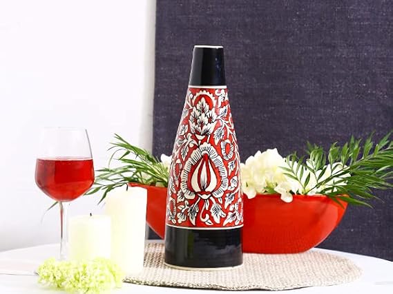 A handmade black and red ceramic Mughal vase with floral patterns, displayed on a table with a red bowl of popcorn, greenery, and a glass of red wine in the background.