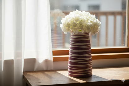 Striped vase with white flowers on a wooden table by a window with sheer curtains.