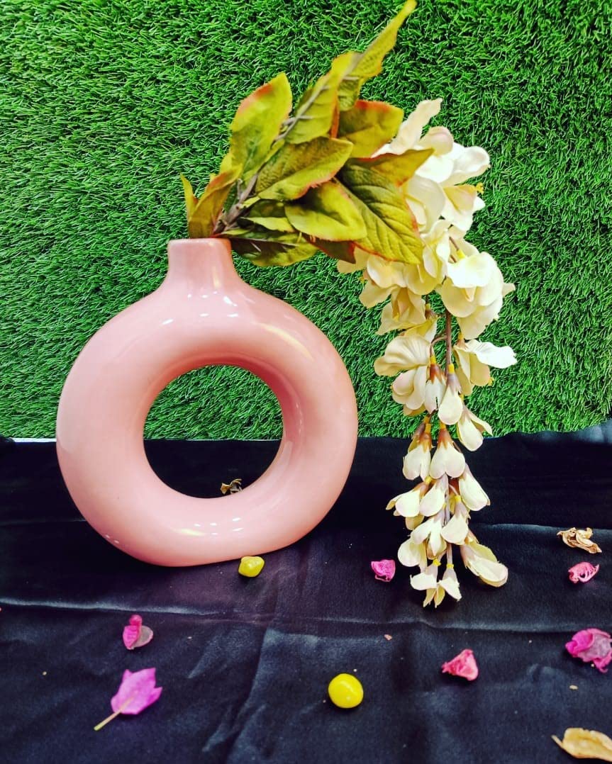 A handmade English pink donut shape ceramic vase with a matte finish, decorated with a floral wreath and placed on a black surface with scattered flower petals around it.