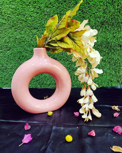 A handmade English pink donut shape ceramic vase with a matte finish, decorated with a floral wreath and placed on a black surface with scattered flower petals around it.