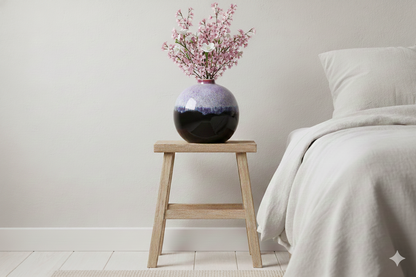 Vase with pink flowers on a wooden stool next to a bed with gray bedding.
