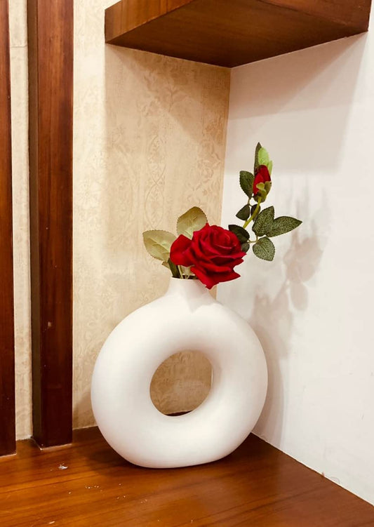 A white matte finish ceramic donut-shaped vase with a few artificial red roses and green leaves placed in it, standing on a wooden surface with a blurred background.