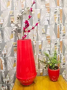 A glossy red cylindrical ceramic vase with a bouquet of pink flowers in it, placed on a wooden surface with a patterned wall in the background.