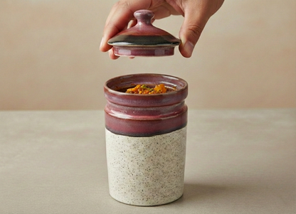 Hand opening a ceramic jar with a red lid on a beige background