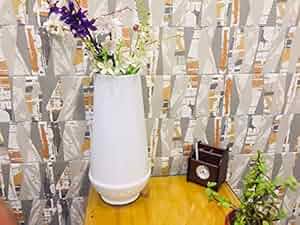 A glossy white ceramic cylindrical vase with a floral arrangement, placed on a side table with a patterned wall and decorative plant in the background.